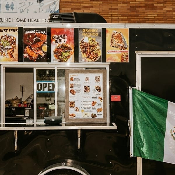 Black food truck with a Mexican flag, menu, and posters of tacos, fries, burritos, and nachos.